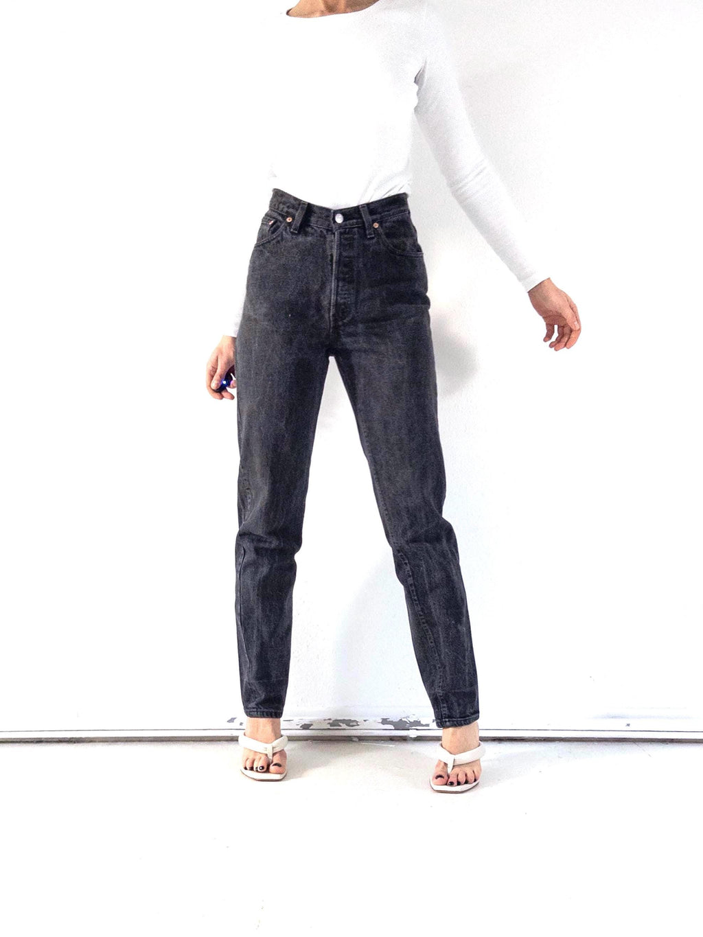 Könnte beinhalten: Schwarze Jeans mit hohem Bund und Knopfleiste. Die Jeans werden mit einem weißen Langarm-Top und weißen Sandalen getragen. Die Jeans haben ein klassisches Fünf-Taschen-Design und eine gerade Beinpassform.