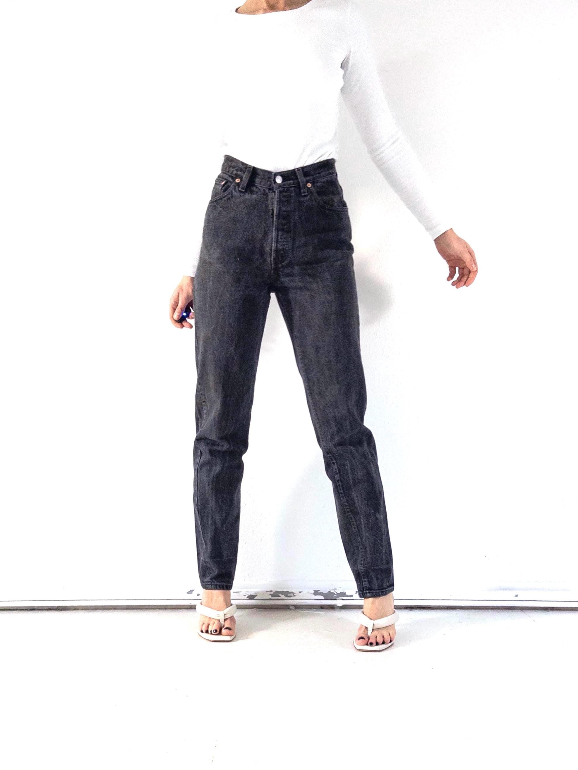 Könnte beinhalten: Schwarze Jeans mit hohem Bund und Knopfleiste. Die Jeans werden mit einem weißen Langarm-Top und weißen Sandalen getragen. Die Jeans haben ein klassisches Fünf-Taschen-Design und eine gerade Beinpassform.