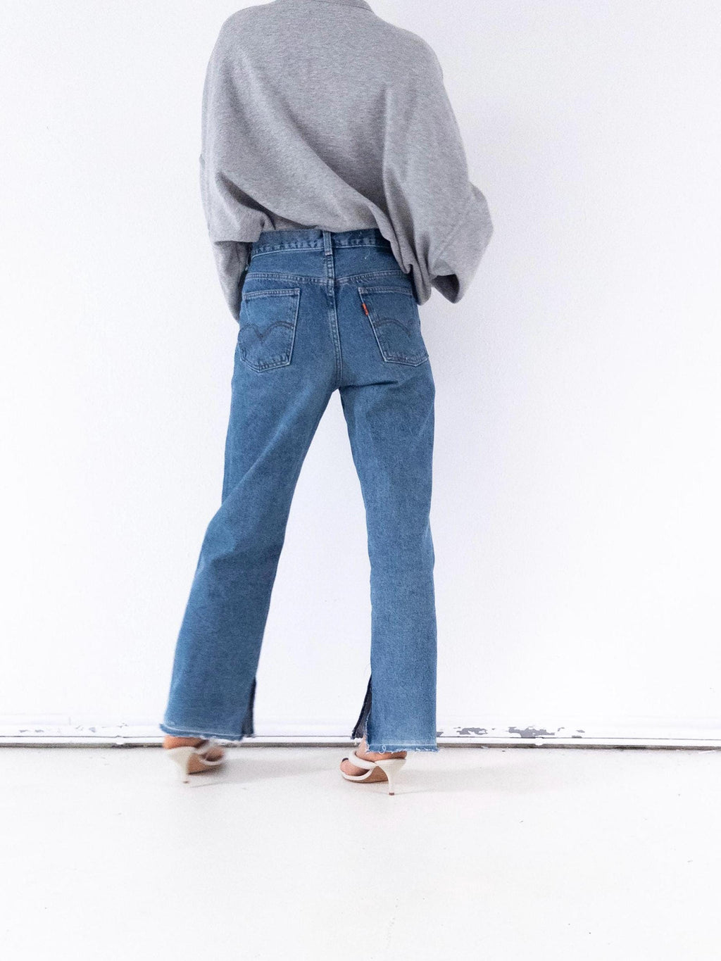 Könnte beinhalten: Eine Person trägt einen grauen Pullover und eine blaue Jeans. Die Jeans hat einen Schlitz am Knöchel und ein rotes Levi's-Etikett auf der Gesäßtasche. Die Person trägt weiße Absätze.