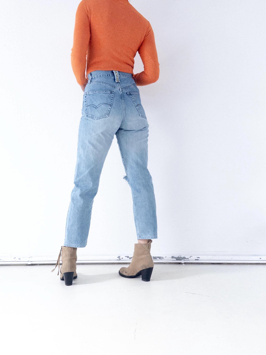 Könnte beinhalten: Hellblaue Jeans mit einem aufgerissenen Knie, kombiniert mit einem orangefarbenen Langarm-Top und beigefarbenen Stiefeletten mit schwarzen Absätzen. Die Jeans hat eine hohe Taille und eine klassische Passform.