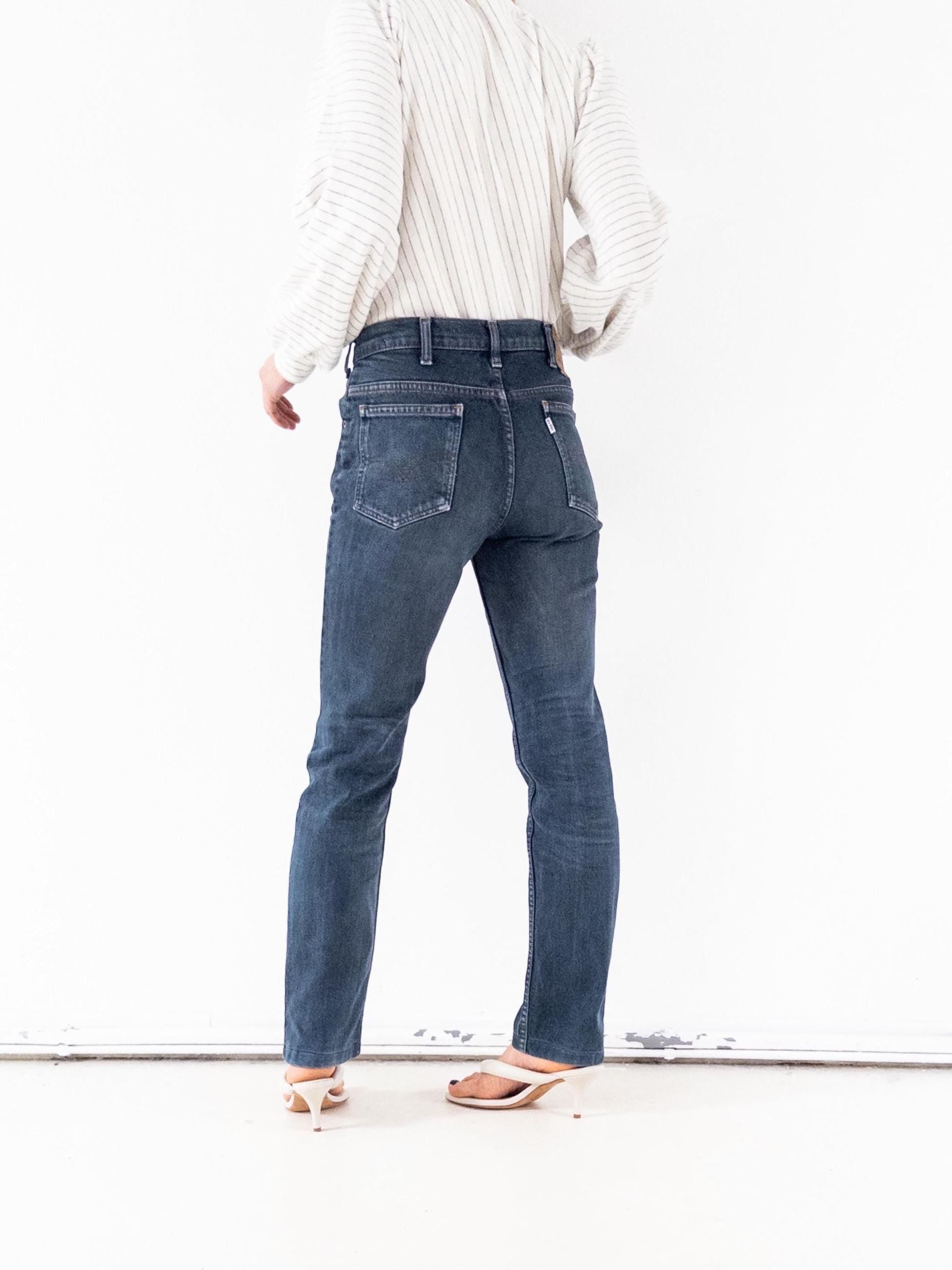 Könnte beinhalten: Dunkelblaue Jeans mit geradem Bein. Die Jeans haben ein klassisches Fünf-Taschen-Design und einen Knopf- und Reißverschluss. Das Model trägt weiße Sandalen mit Absatz und ein gestreiftes Button-Down-Hemd.