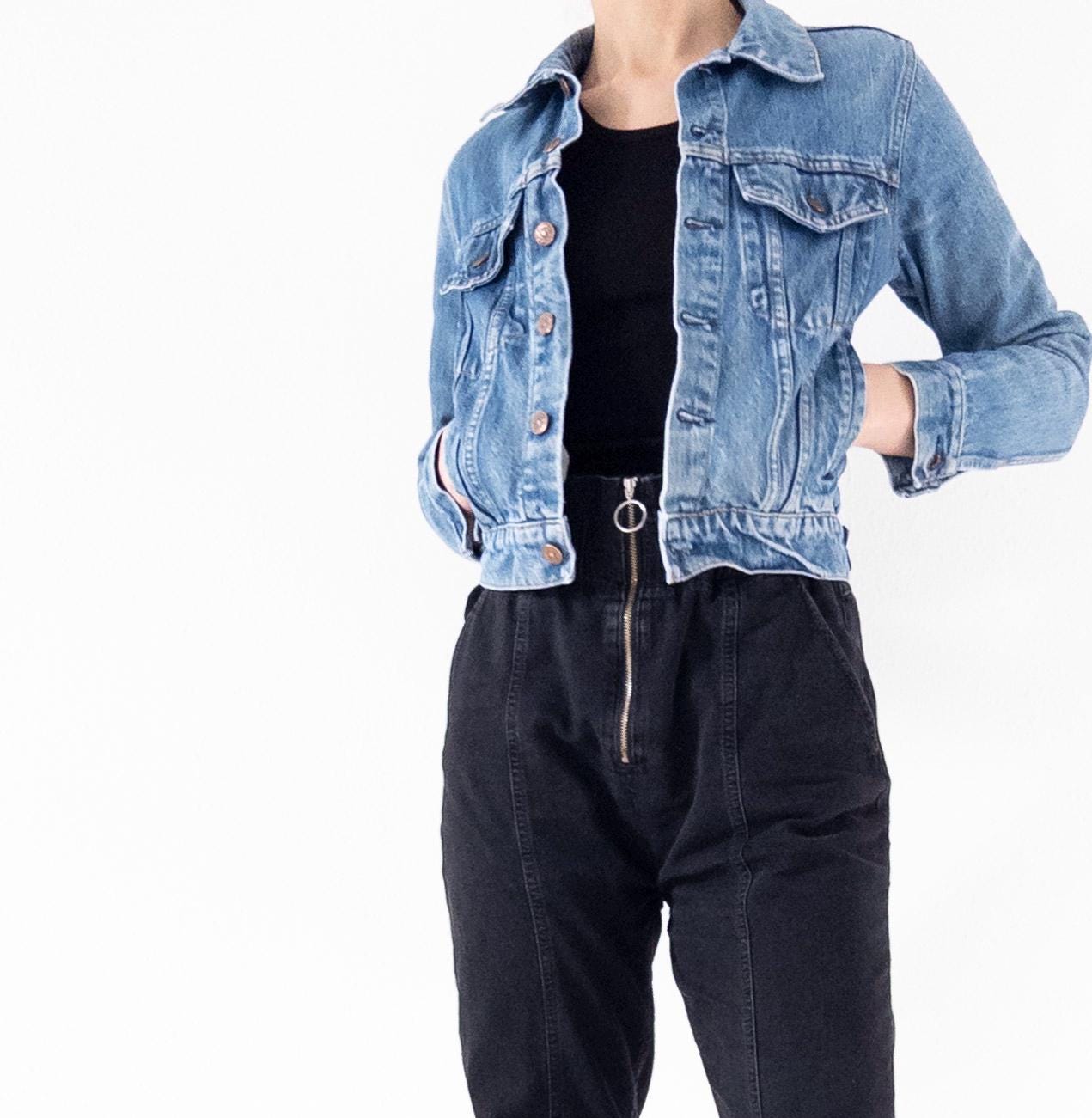 Könnte beinhalten: Eine hellblaue Jeansjacke mit einem schwarzen Oberteil darunter. Die Jacke hat einen Knopfverschluss und zwei Fronttaschen.