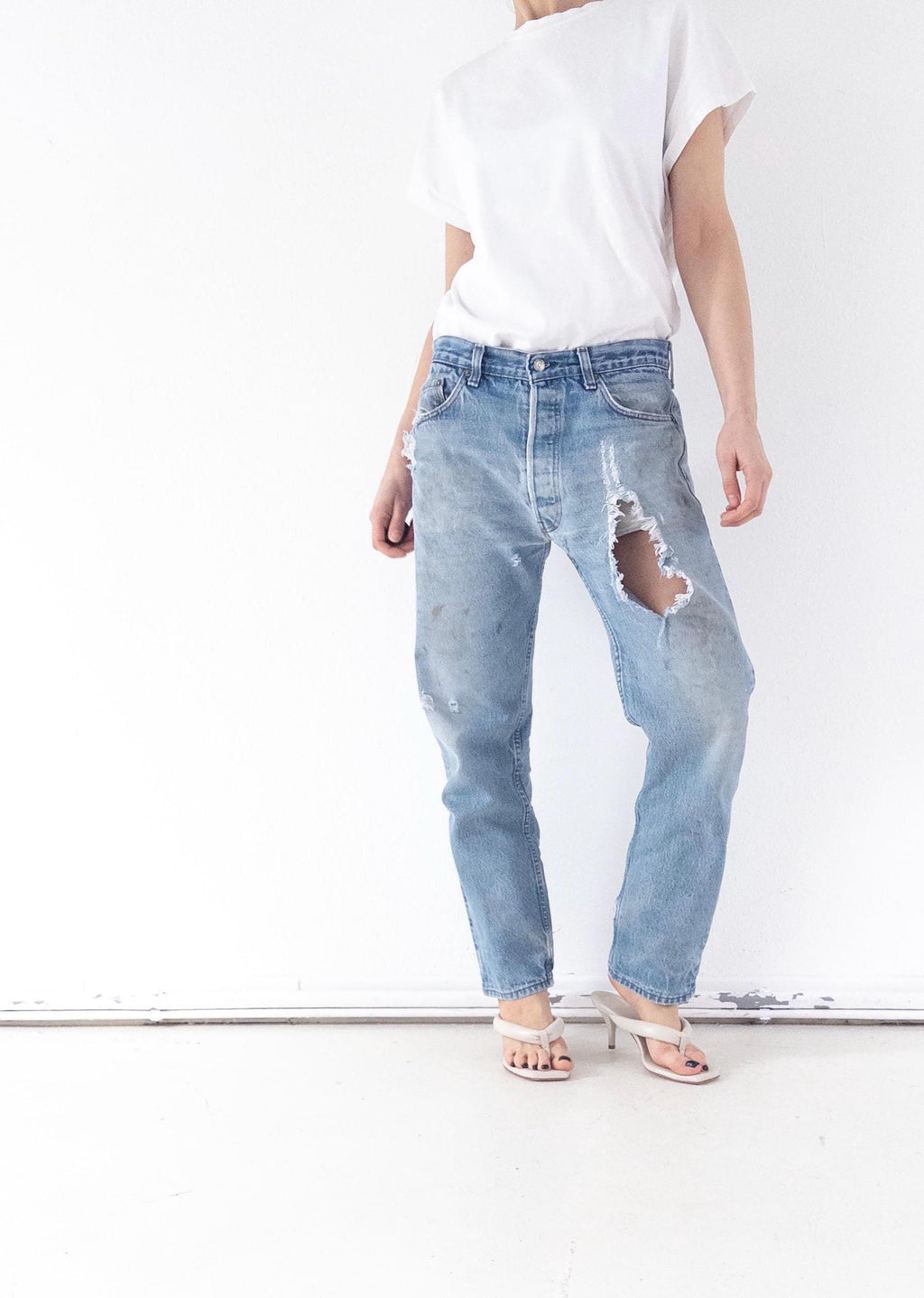 Könnte beinhalten: Hellblaue Jeans mit Distressed-Details, einschließlich großer Risse an den Beinen. Die Jeans werden mit einem weißen T-Shirt und weißen Sandalen mit Absatz kombiniert. Der Gesamtstil ist lässig und vintage.