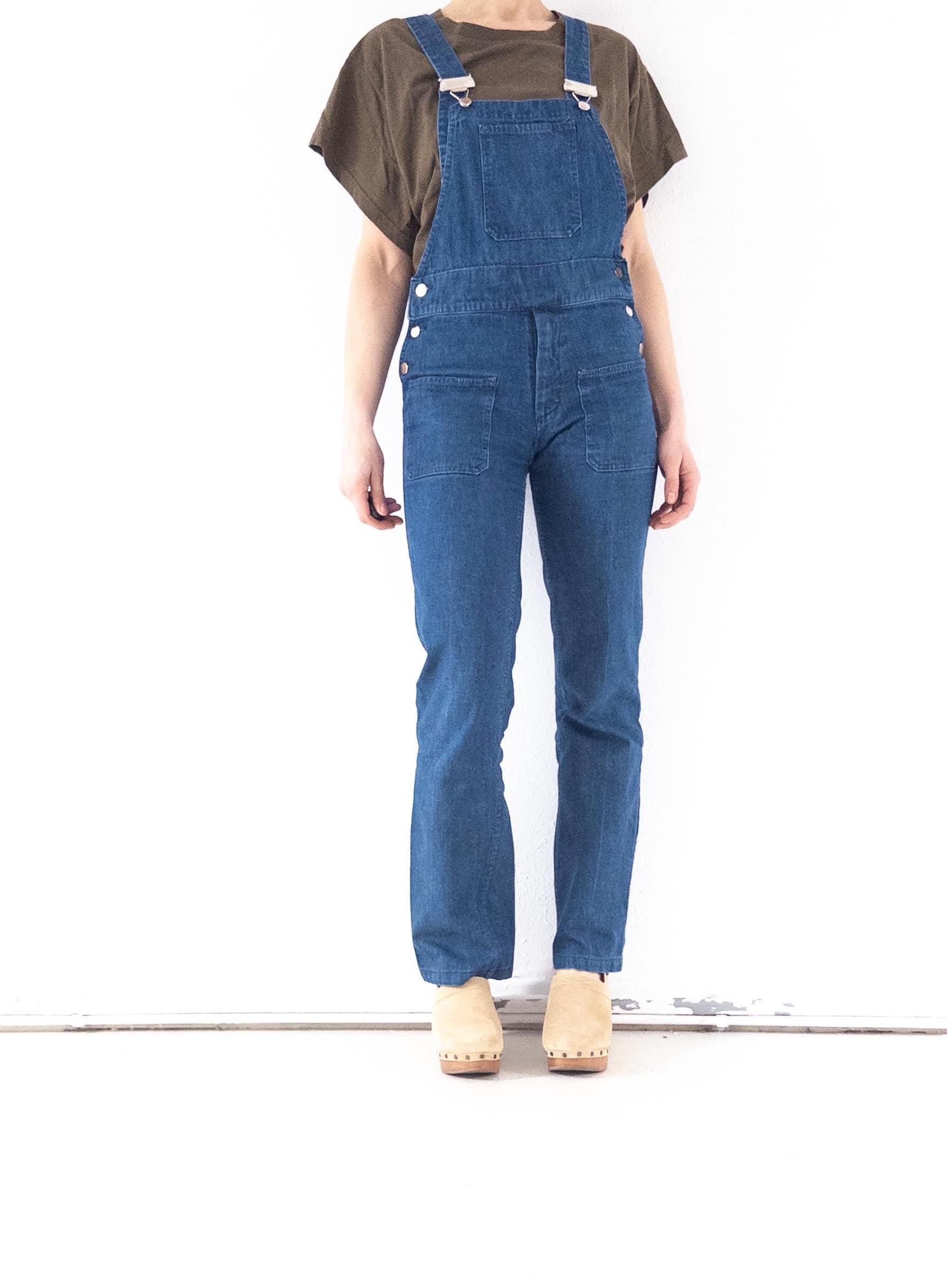 Könnte beinhalten: Blaue Jeans-Latzhose mit verstellbaren Trägern und Taschen. Die Latzhose hat silberfarbene Details und wird mit einem olivgrünen Kurzarm-T-Shirt und beigen Clogs getragen.