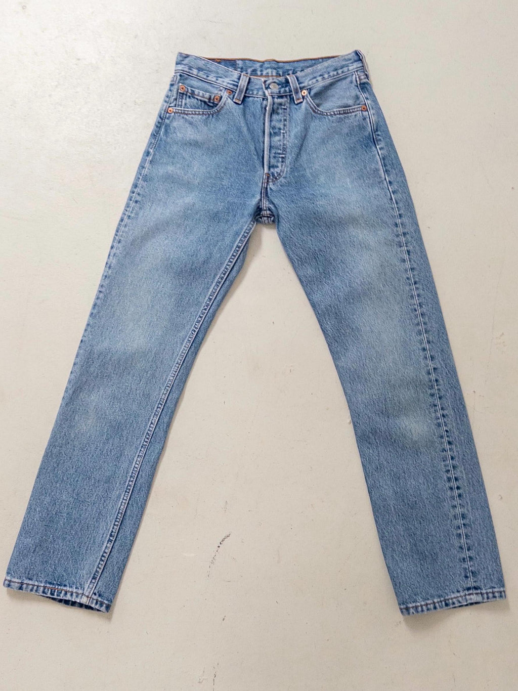 Hellblaue Vintage Levis 501 W26 L30 Made in USA flach ausgelegt. Vollständige Vorderansicht mit charakteristischer Waschung und klassischem Straight-Leg-Schnitt.