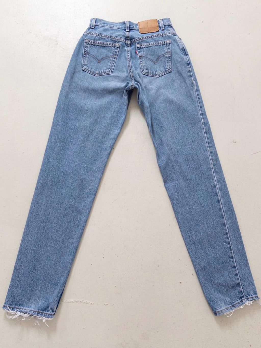 Flatlay Rückseite der Vintage Levis 550 High Waist Jeans W26 L34 in light to mid Blue stonewash. Liegendes Produktfoto mit Details wie ausgefransten Taschen & Saum