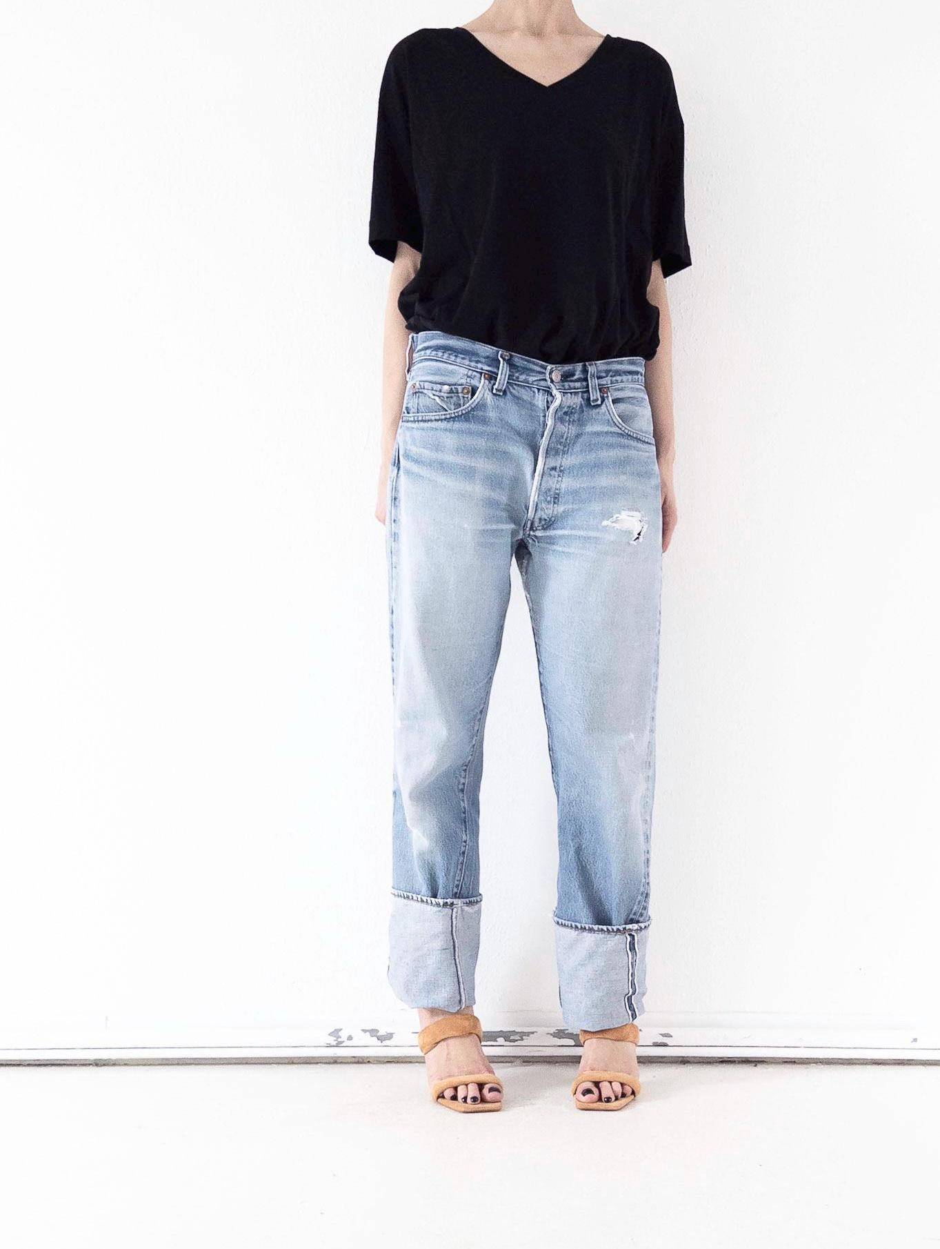 Hellblaue Levi’s 501 Selvedge Jeans in der Frontansicht. Vintage One of a Kind Unikat mit dezenter Grunge-Waschung, Distressed-Details am Oberschenkel und Umschlag. Gestylt mit lockerem schwarzem V-Shirt und Sandaletten von girlstalkvintage.