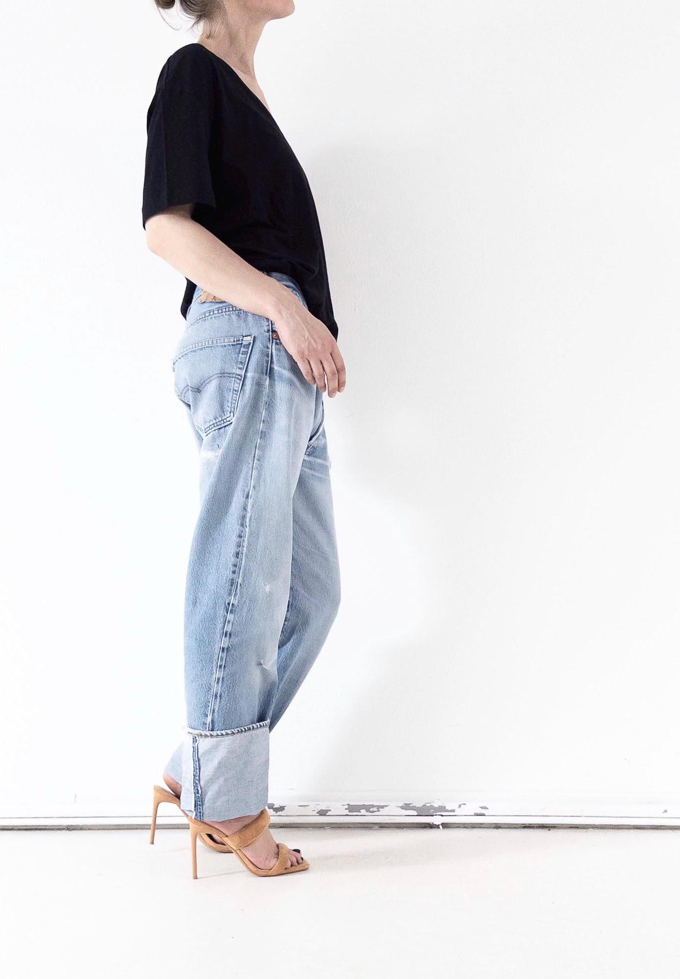 Seitenansicht derlight wash unisex Vintage Levi’s 501 Selvedge Jeans im Grunge Distressed-Look mit breitem Umschlag und charakteristischer Webkante.  One of a kind Styling mit schwarzem Top und filigranen High Heels. Authentischer 80s Thrashed Denim im loose Boyfriend Look von girlstalkvintage.