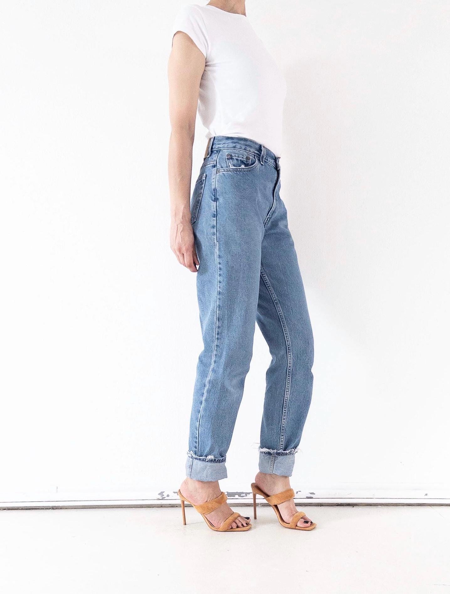 Seitwnsicht der Vintage Levis 550 High Waist Jeans W26 L34. Zeigt den Bund, die vorderen Taschen & den Tapered-Schnitt, gestylt mit weißen 70s Top und Highheels, die den ausgefransten Saum betonen.